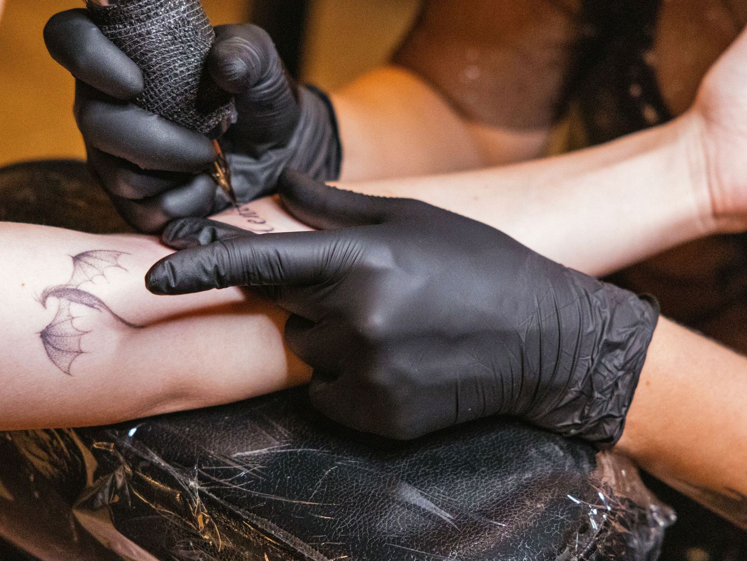 Tattoo artist wearing black gloves carefully inking a delicate floral design on a client's forearm.