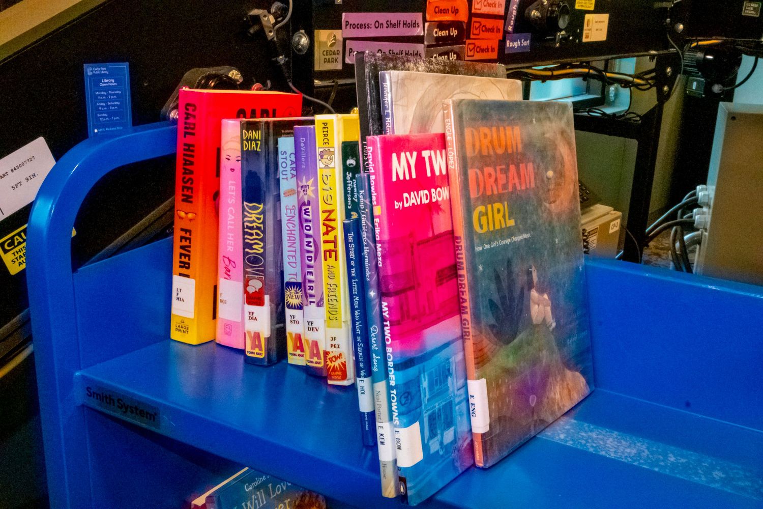 Colorful books displayed on a blue library cart, including My Two Dream Girl by David Kap.