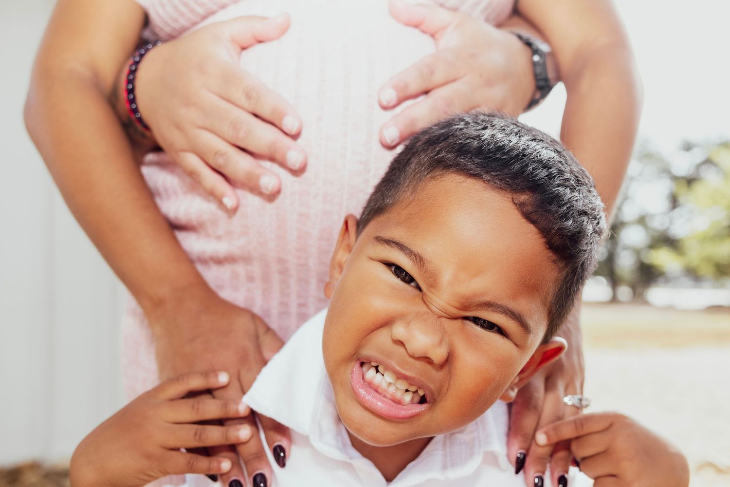 A young boy smiles in front of a pregnant woman's belly, touching her bump in a sweet maternity photo.
