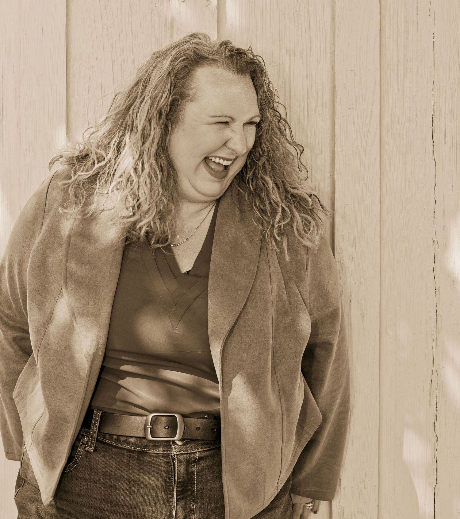 A woman with curly hair laughs joyfully in a sepia-toned photo, wearing a blazer and jeans against a wooden wall.