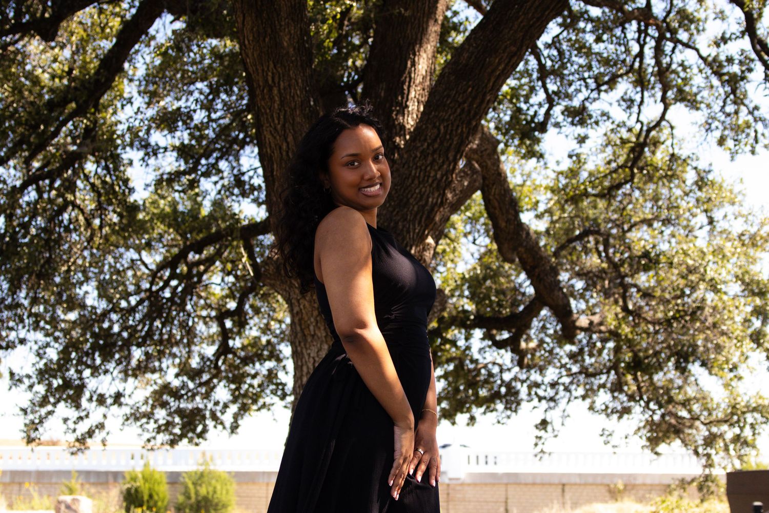 A smiling woman in a black dress poses gracefully beside a large oak tree outdoors.