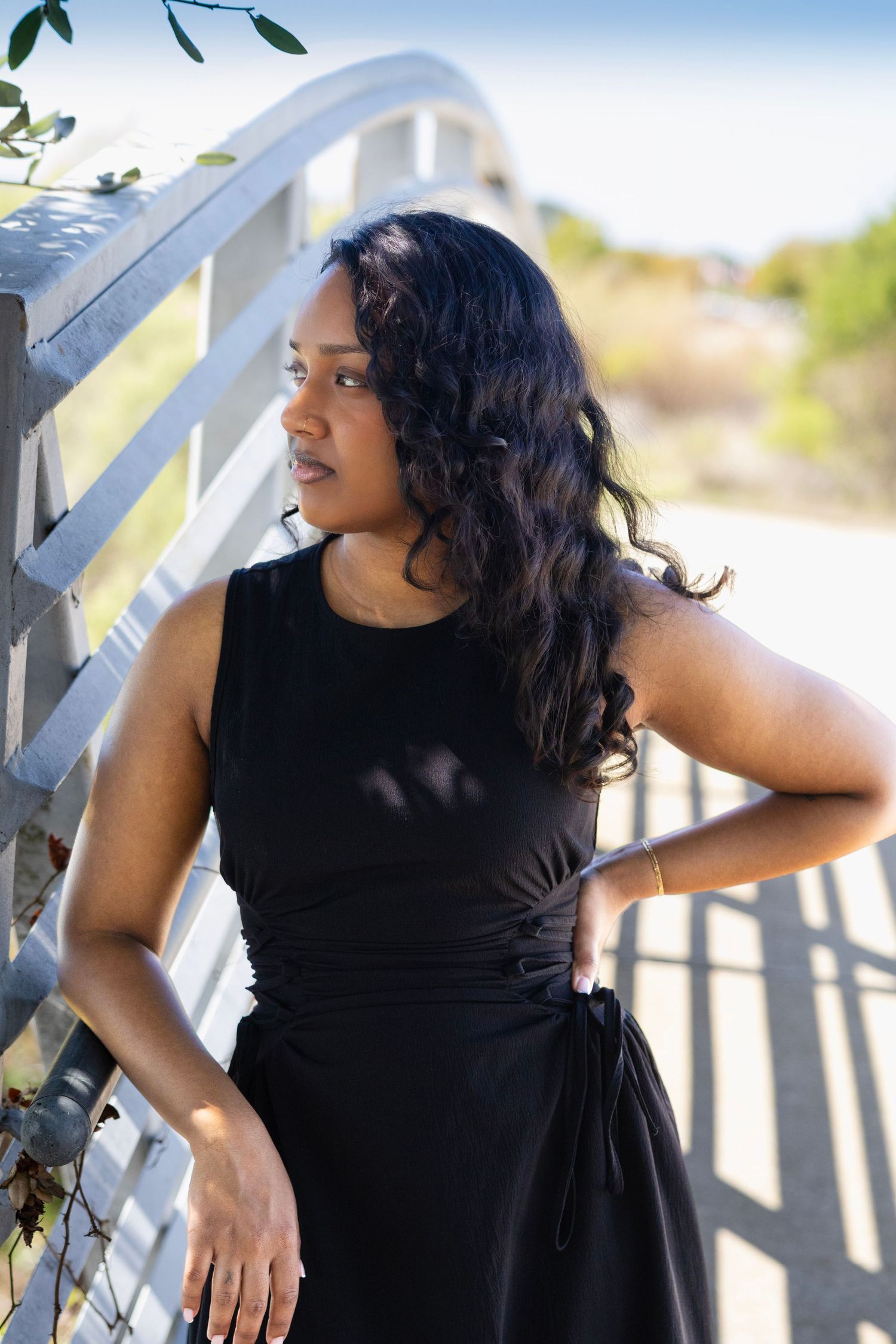 A Black woman in a black sleeveless dress leans against a white wooden bridge railing outdoors.