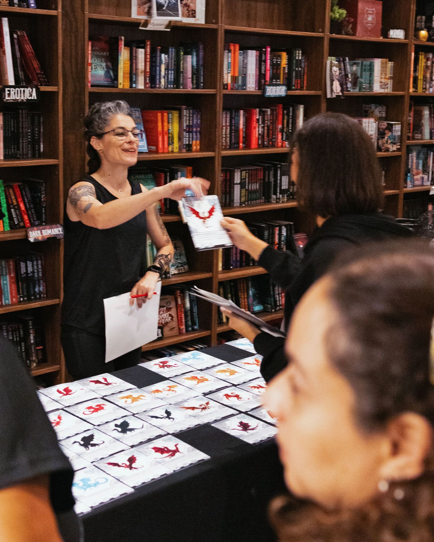 A tattooed woman with gray hair signs books at a bookstore table displaying illustrated cards for fans.
