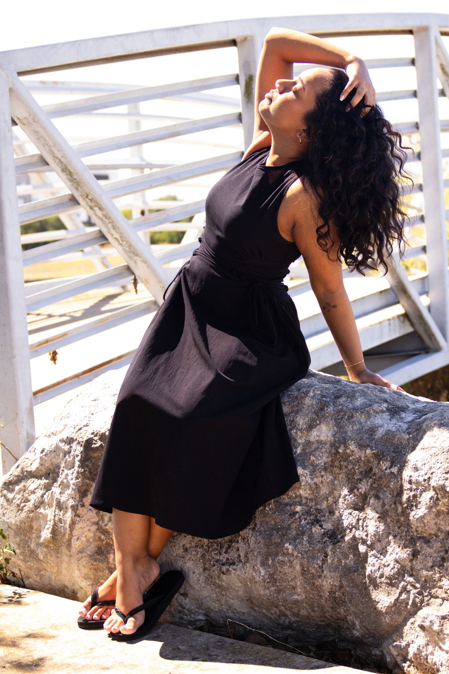Young Black woman in black midi dress posing on rocks near white bridge railing on sunny day.
