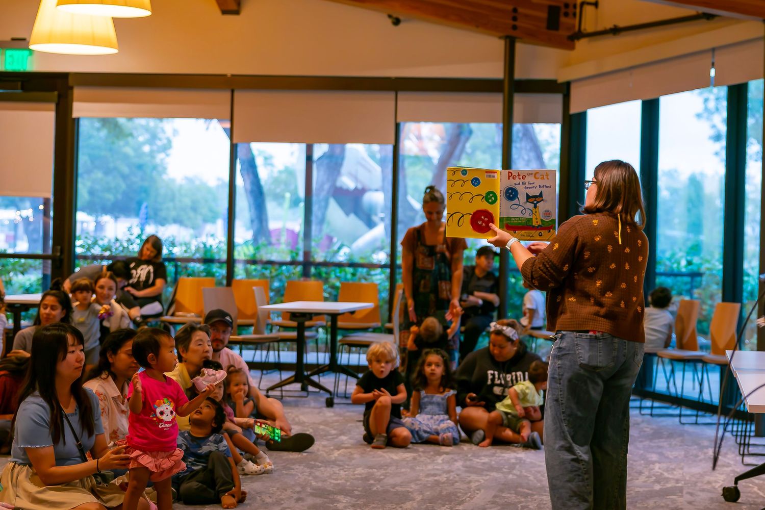 A teacher reads a colorful picture book to a group of children and parents seated on the floor in a modern library.
