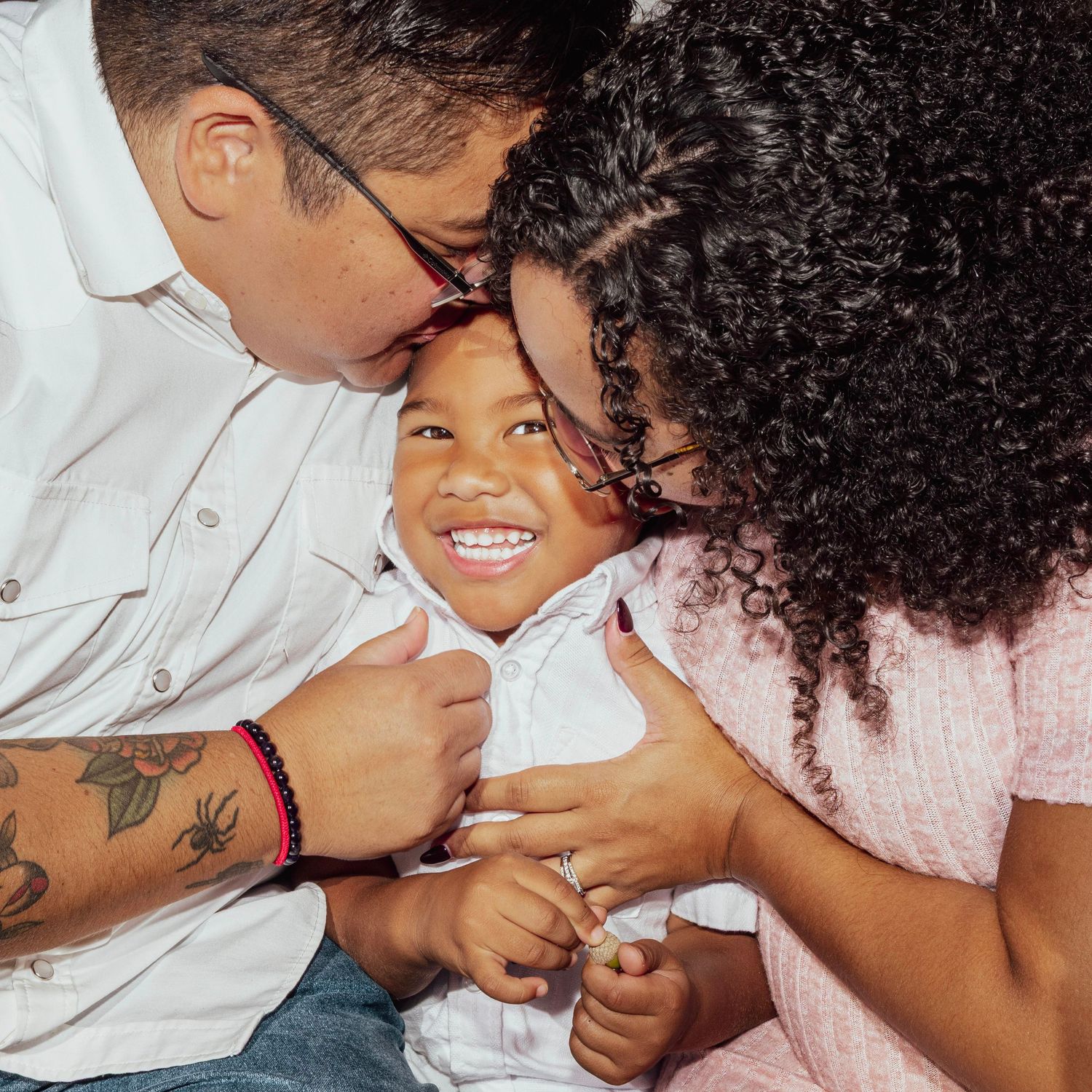 A joyful family moment with parents kissing their smiling toddler, dad showing a tattoo sleeve, mom with curly hair.