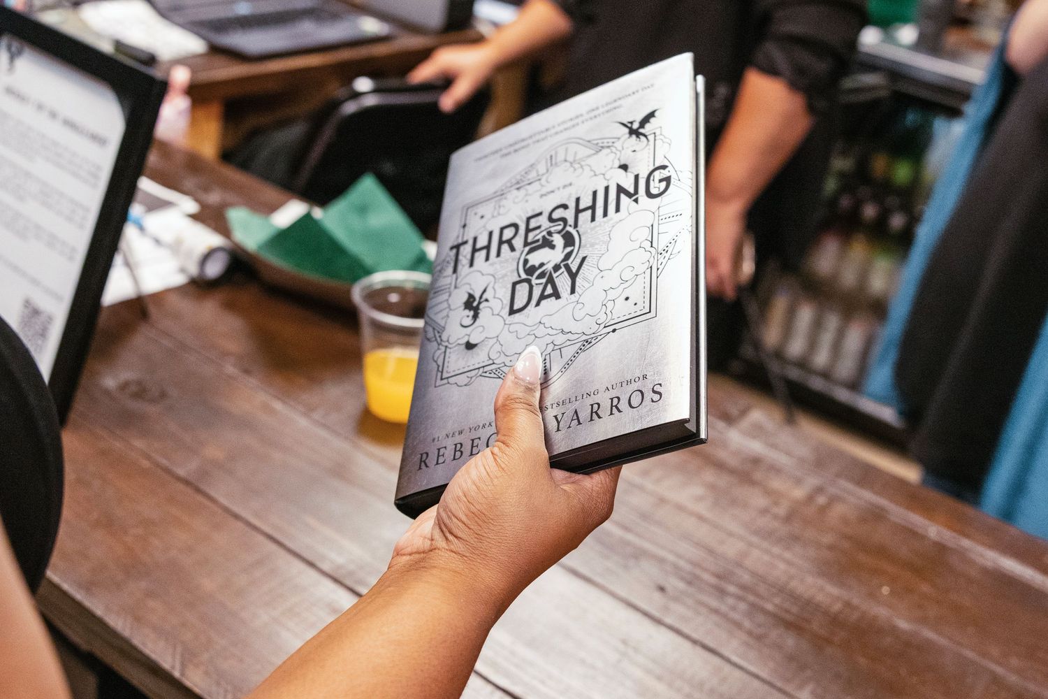 Hand holding a copy of Threshing of Days book by Rebecca Yarros at a wooden table.