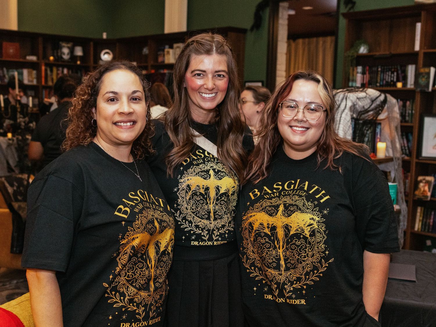 Three women wearing matching Basgiath black and gold t-shirts smile together inside a bookstore.