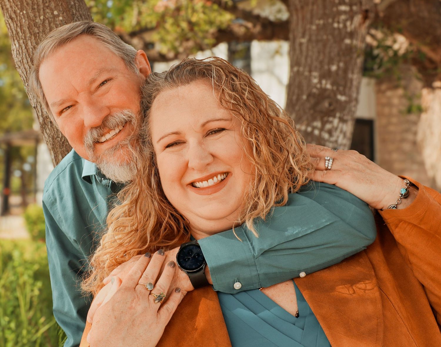 Happy couple embracing outdoors near trees, man hugging woman from behind, both smiling joyfully.