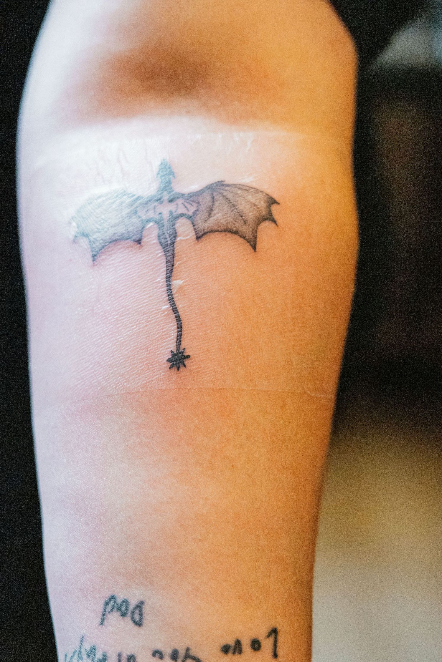 Small dragon tattoo with bat wings on an arm, featuring fine line black ink detail.