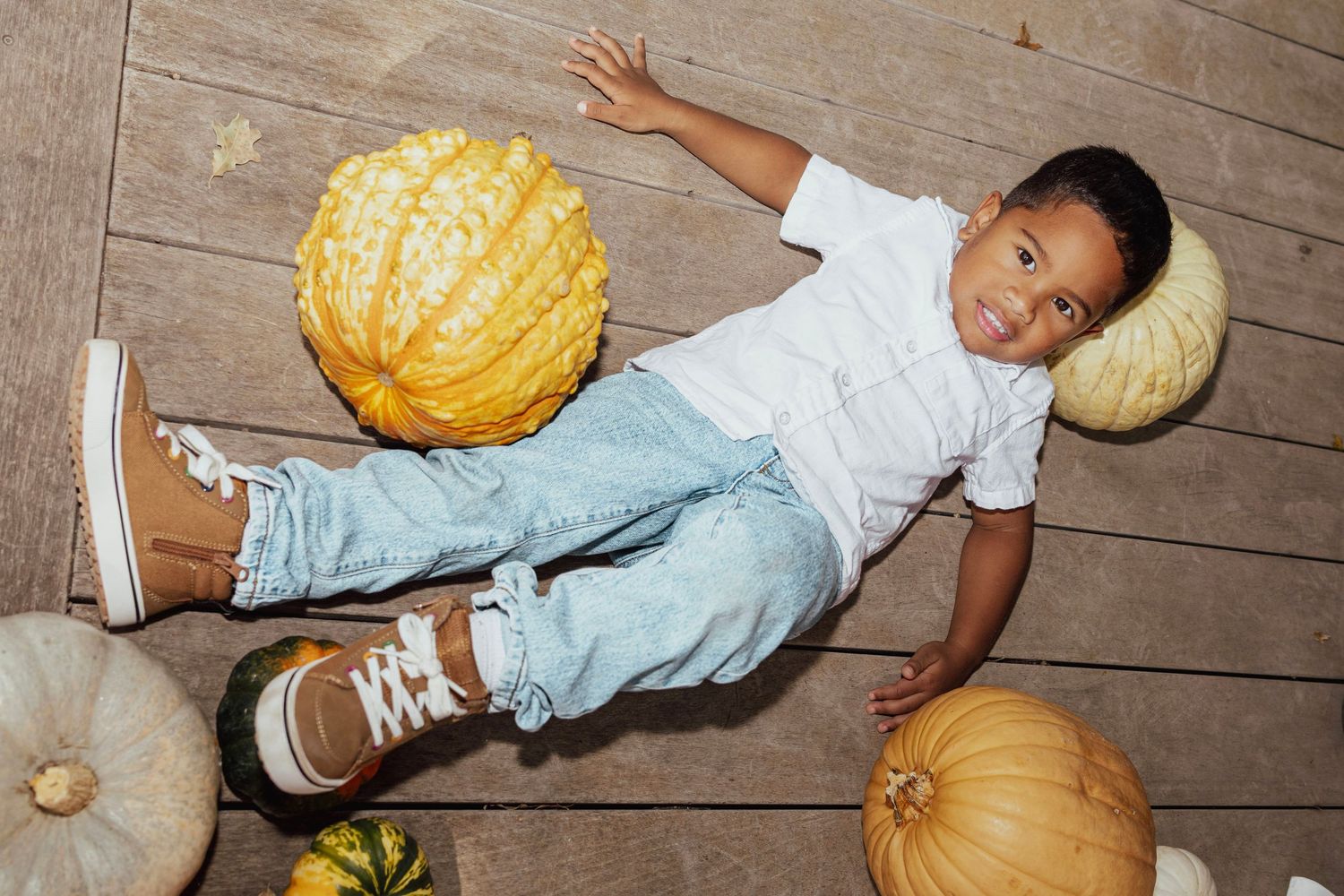 A joyful child lying among colorful pumpkins on a wooden floor, wearing jeans and brown sneakers during fall.