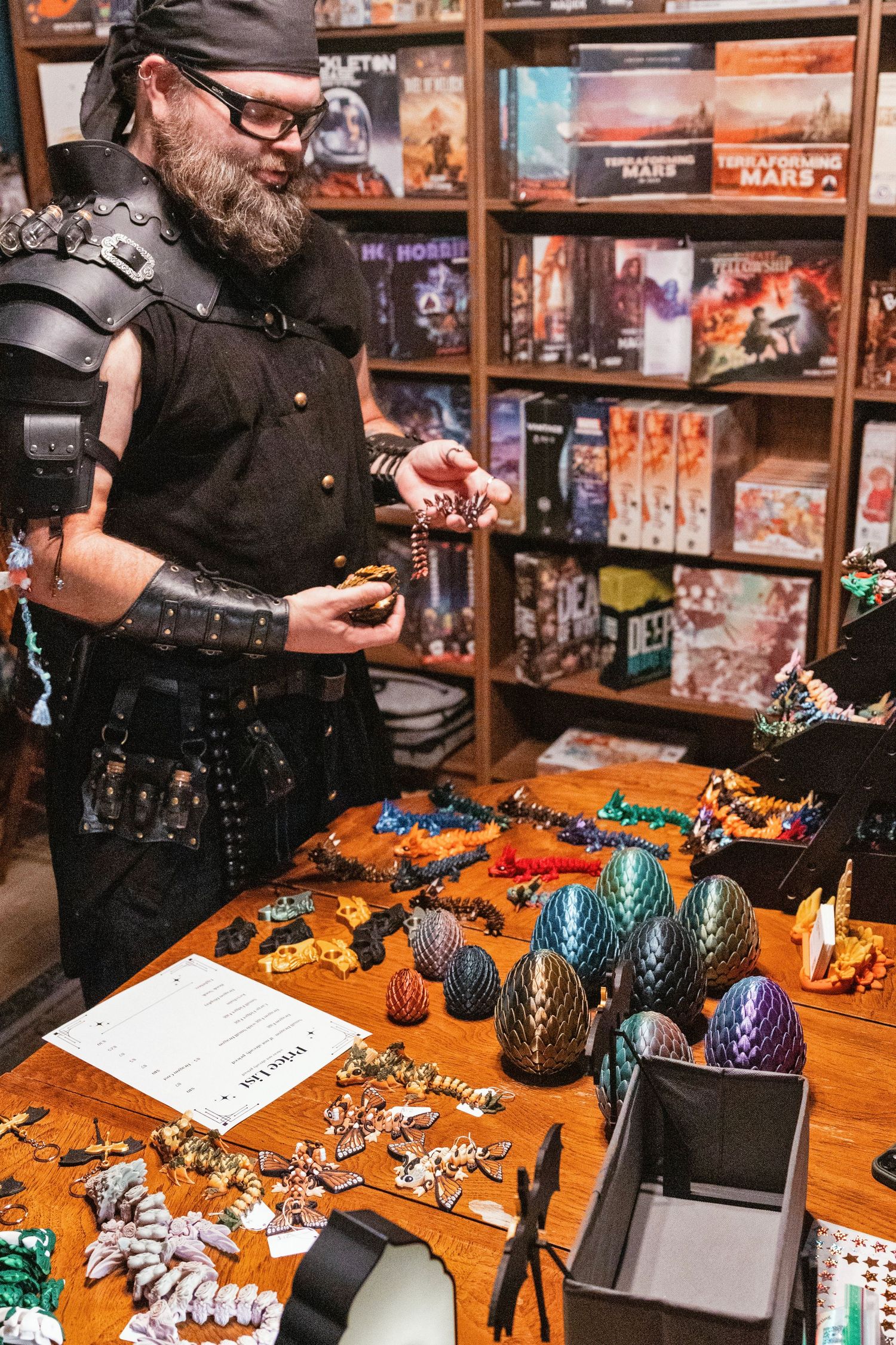 Bearded man in medieval armor examines dragon eggs and fantasy figurines at a game store vendor table.