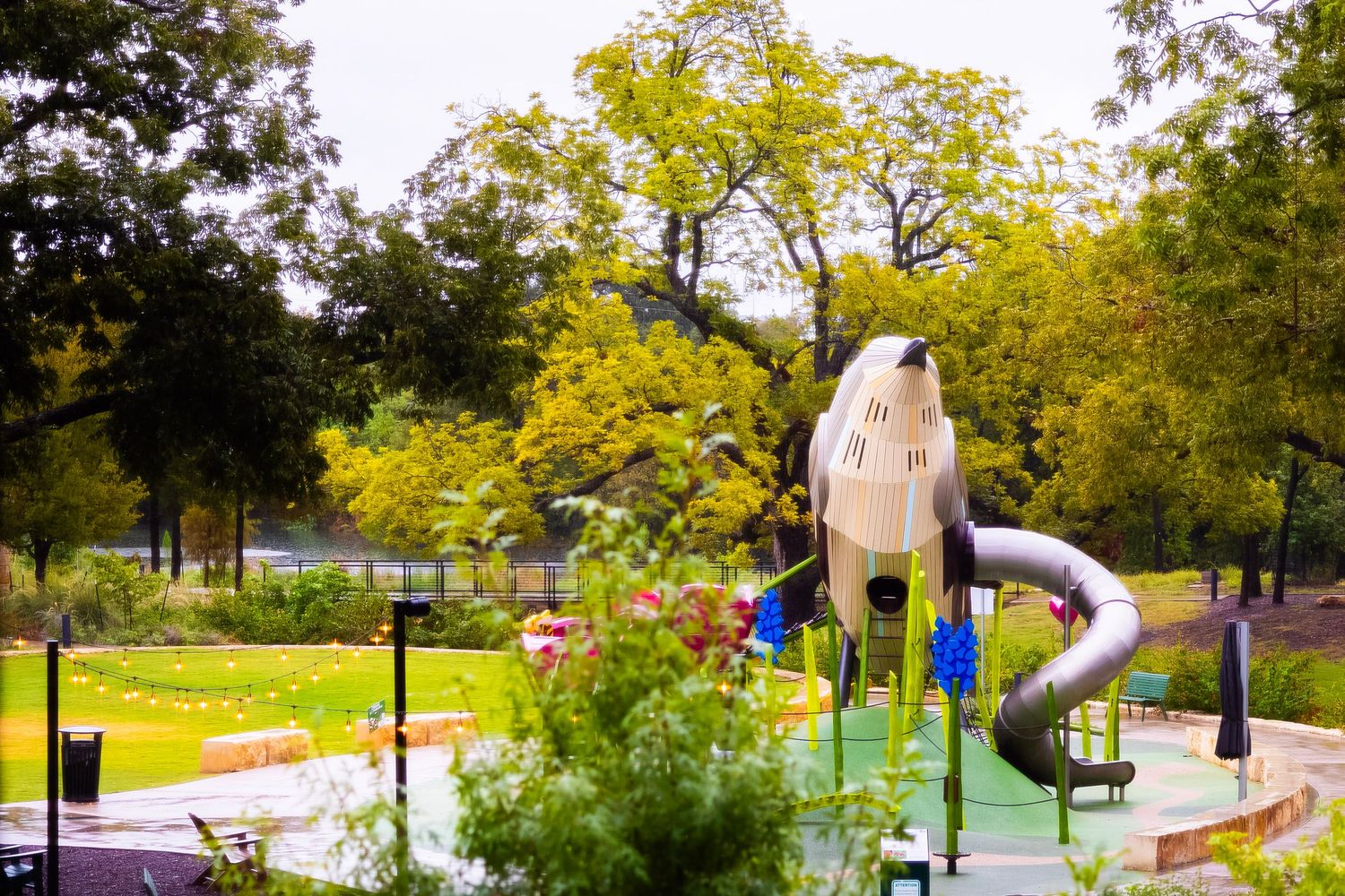 Colorful playground with rocket slide structure surrounded by lush green trees in a scenic park setting.