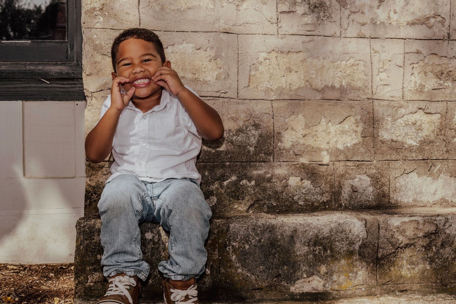 Smiling young boy sitting on stone steps, hands on cheeks, wearing white shirt and jeans.