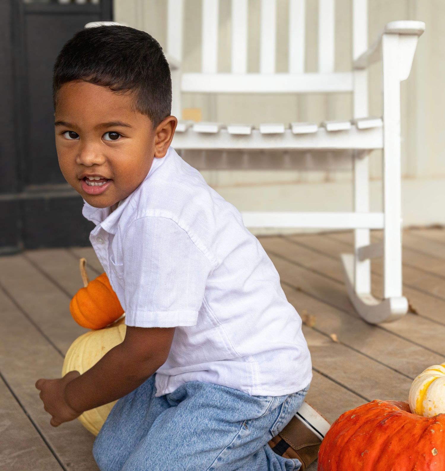 A young boy poses with colorful pumpkins on a porch next to a white rocking chair in a fall photo.