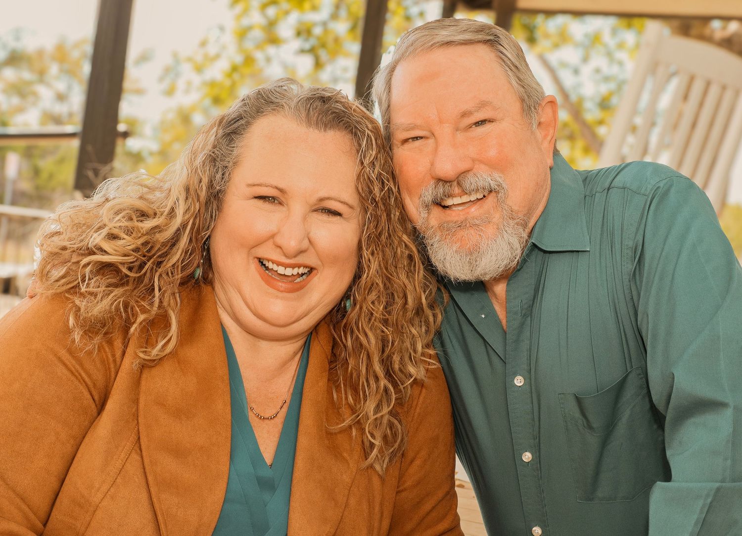 A smiling middle-aged couple posing closely together outdoors, woman with curly hair in brown jacket, man with beard in teal shirt.
