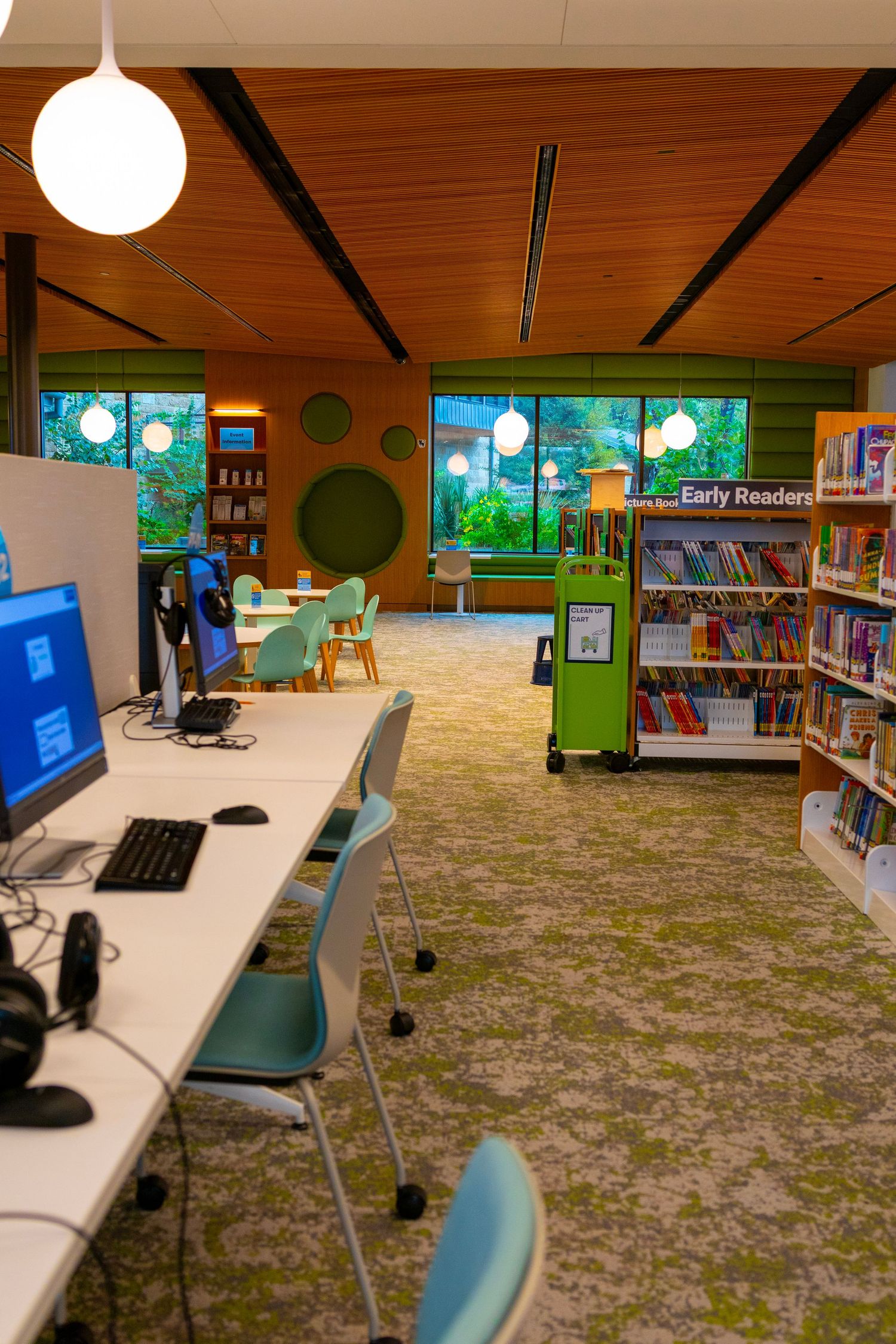 Bright children's library with computer stations, colorful bookshelves, green decor, and an Early Readers section under a warm wood ceiling.