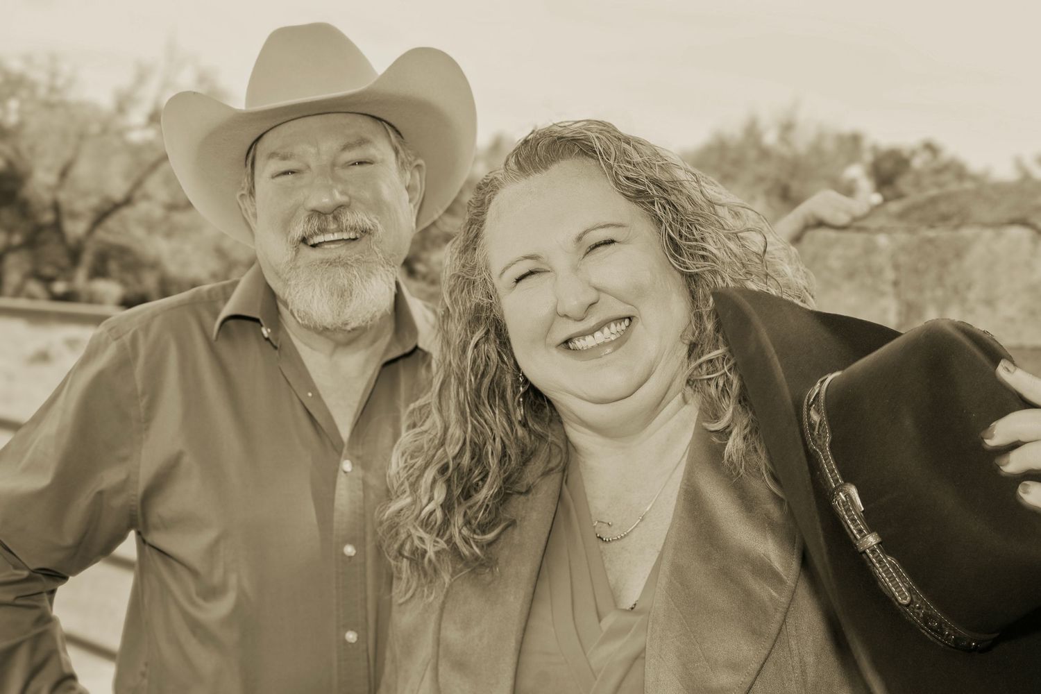 Smiling man in cowboy hat and woman with curly hair pose together in a warm sepia-toned outdoor portrait.