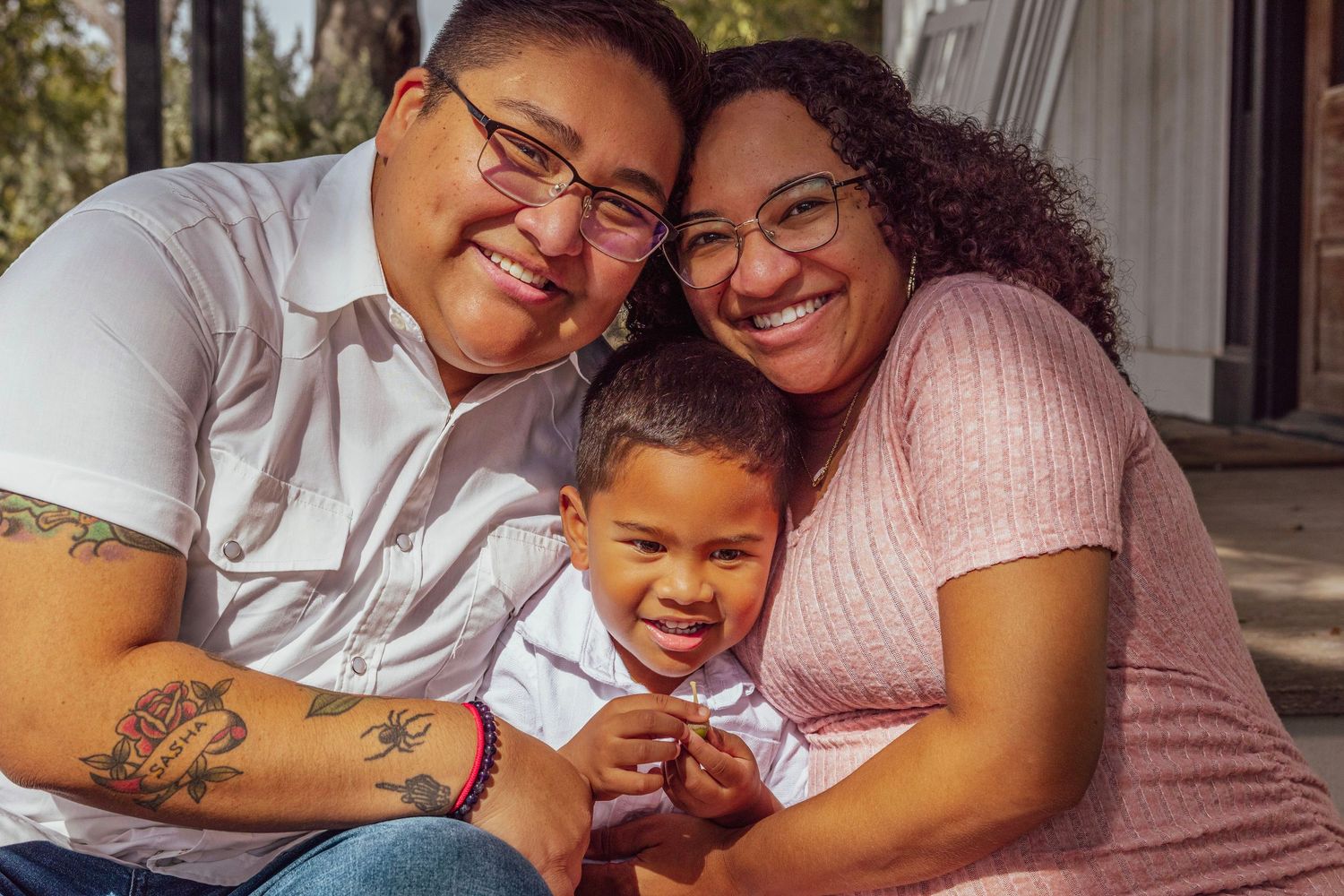 Happy family portrait of two mothers and their young son smiling together on an outdoor porch step.