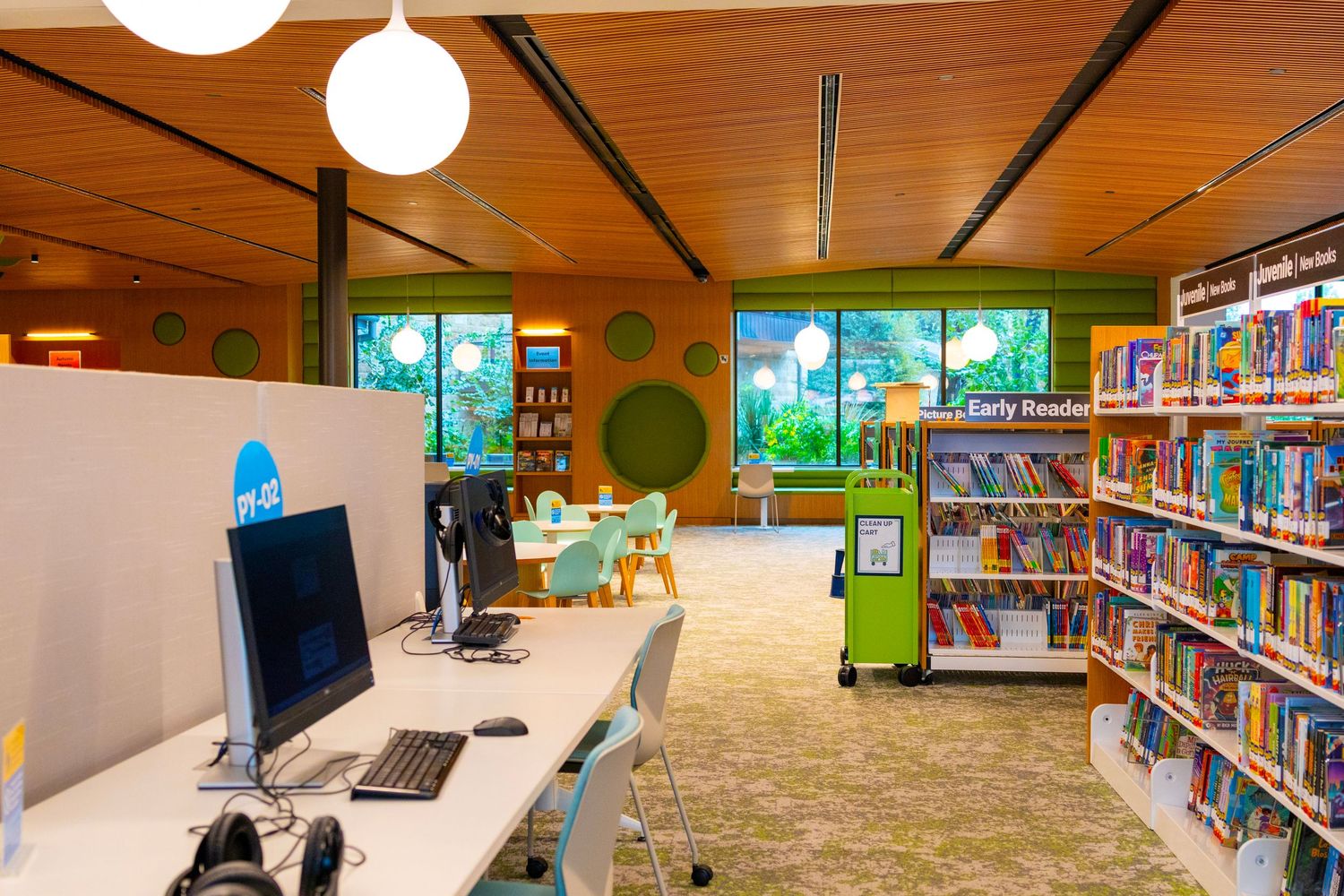 Bright children's library section with computers, colorful bookshelves, and early readers area under warm wooden ceiling.