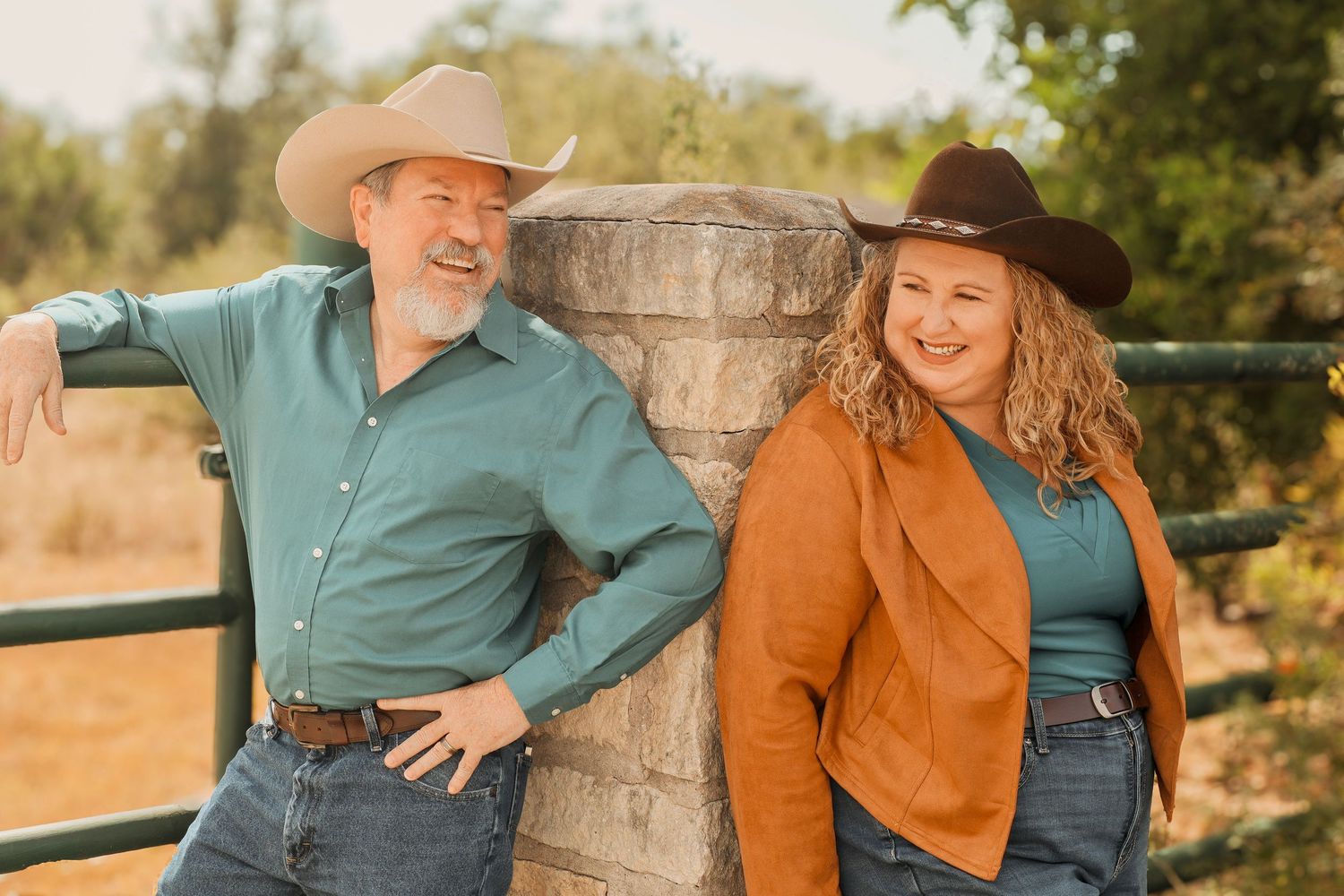 A man and woman in cowboy hats lean against a wooden post by a ranch fence in a sunny outdoor setting.