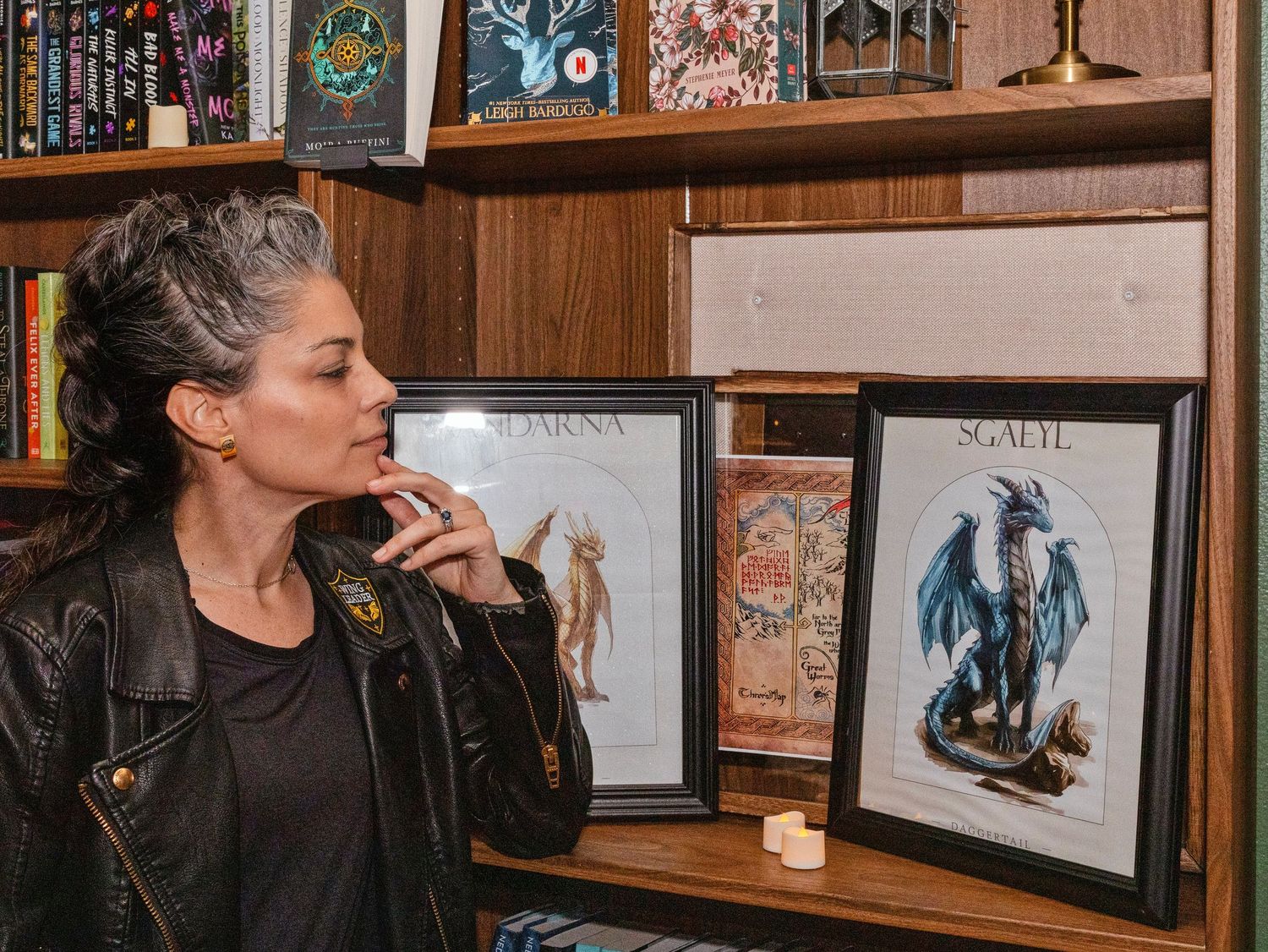 Woman in leather jacket examining framed dragon artwork on wooden bookshelf filled with books and collectibles.