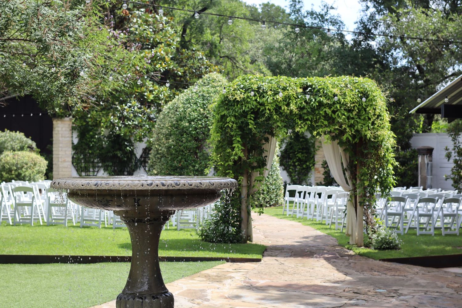 Photo by Lexy Hiam Elegant outdoor wedding venue with a stone fountain, ivy-covered arch, and white chairs on a garden path.