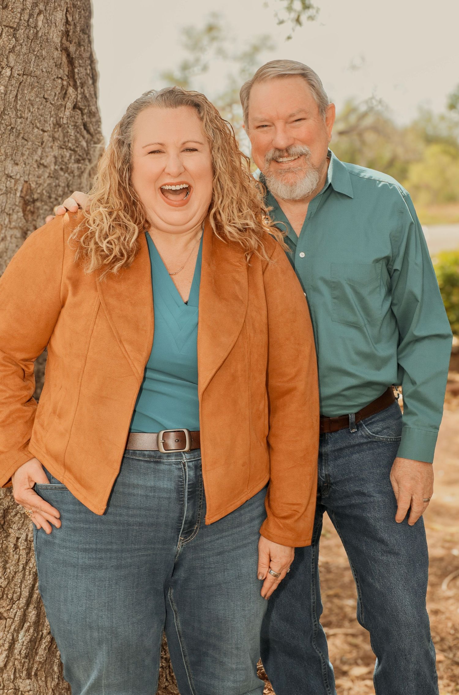 Smiling couple posing outdoors near a tree, woman in orange jacket and man in teal shirt, both wearing jeans.