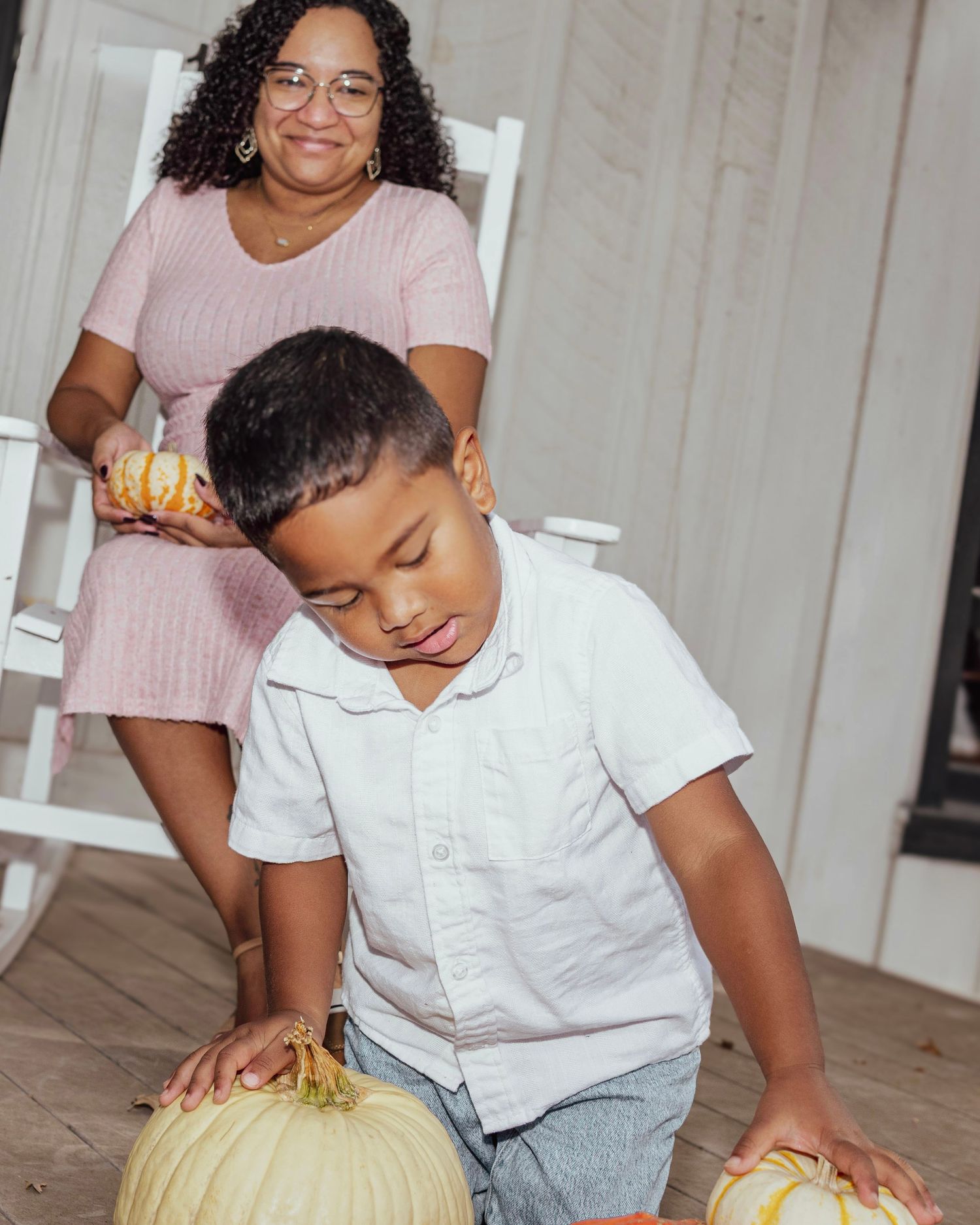 A grandmother and young boy enjoy fall pumpkins on a porch, with the woman holding a small gourd in a pink dress.