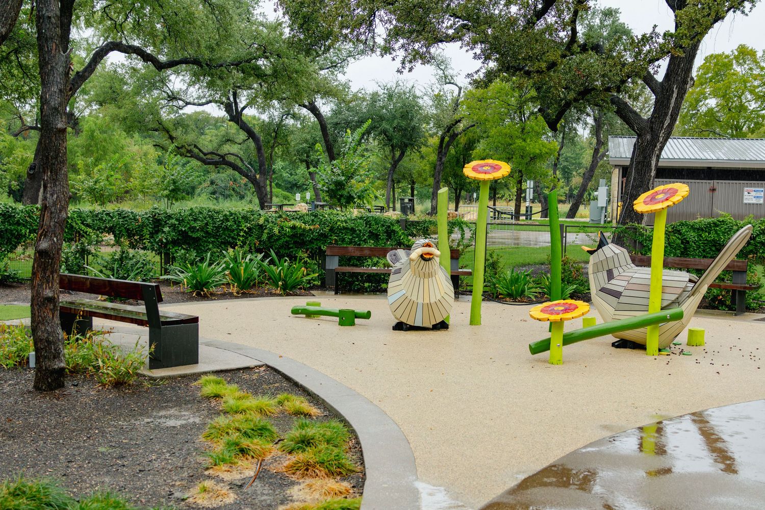 Children's playground with nature-themed equipment, sandy surface, and lush green trees in a park setting.