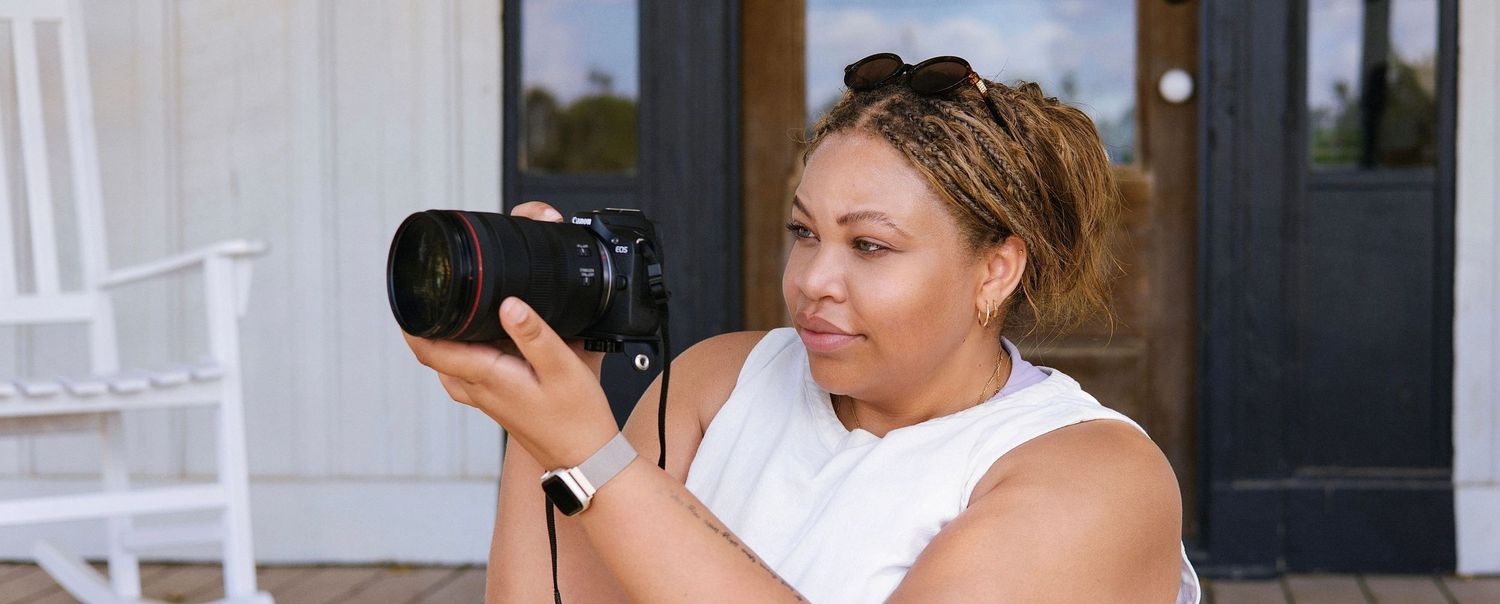A woman in a white sleeveless top examines a professional DSLR camera outdoors near a white porch.