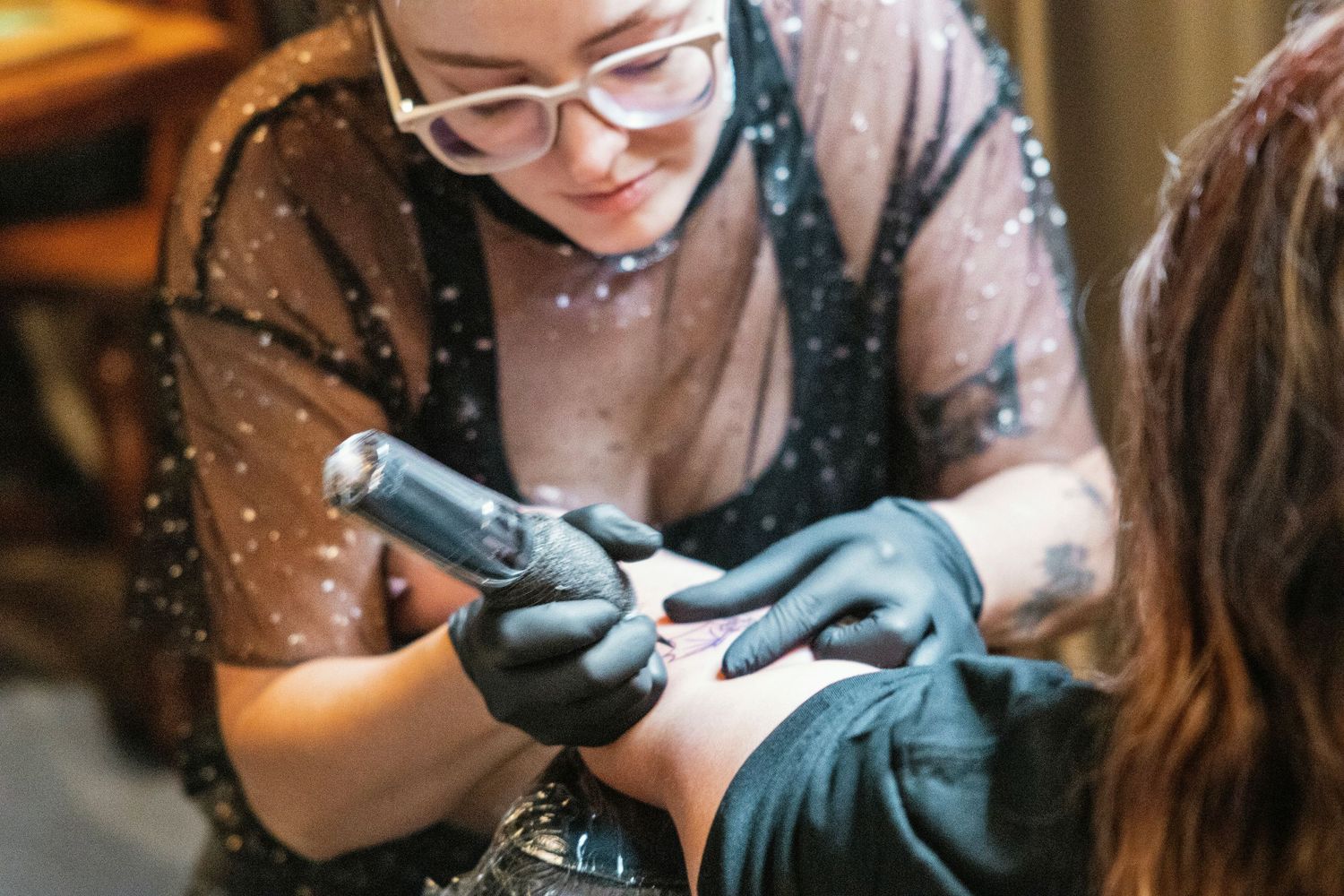 Female tattoo artist wearing black gloves and glasses carefully applies a tattoo design to a client's hand.