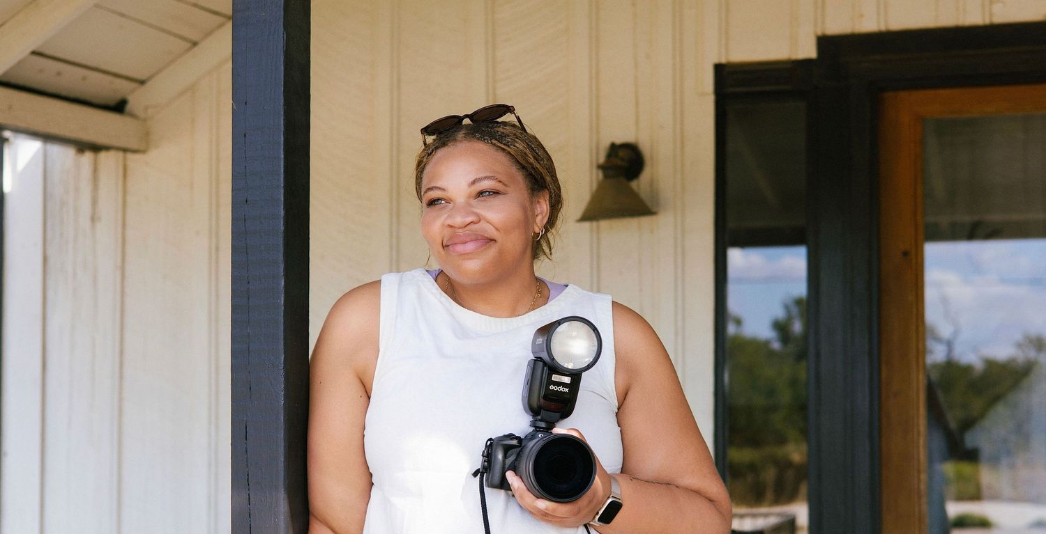Smiling woman holding a professional camera on a porch, wearing a white sleeveless top with sunglasses on her head.