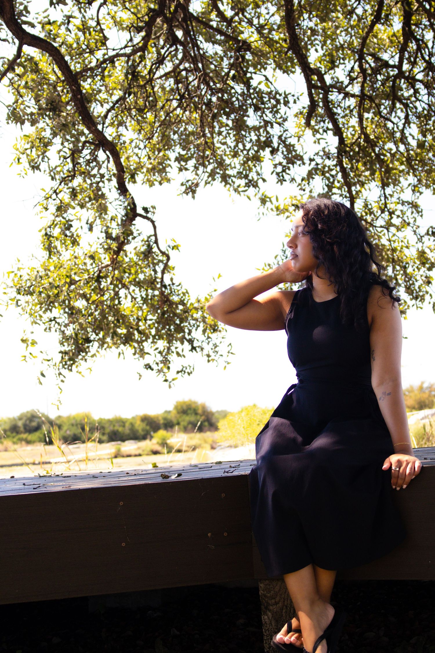 Woman in black dress sitting on wooden ledge under oak tree, smiling in sunlit outdoor setting.