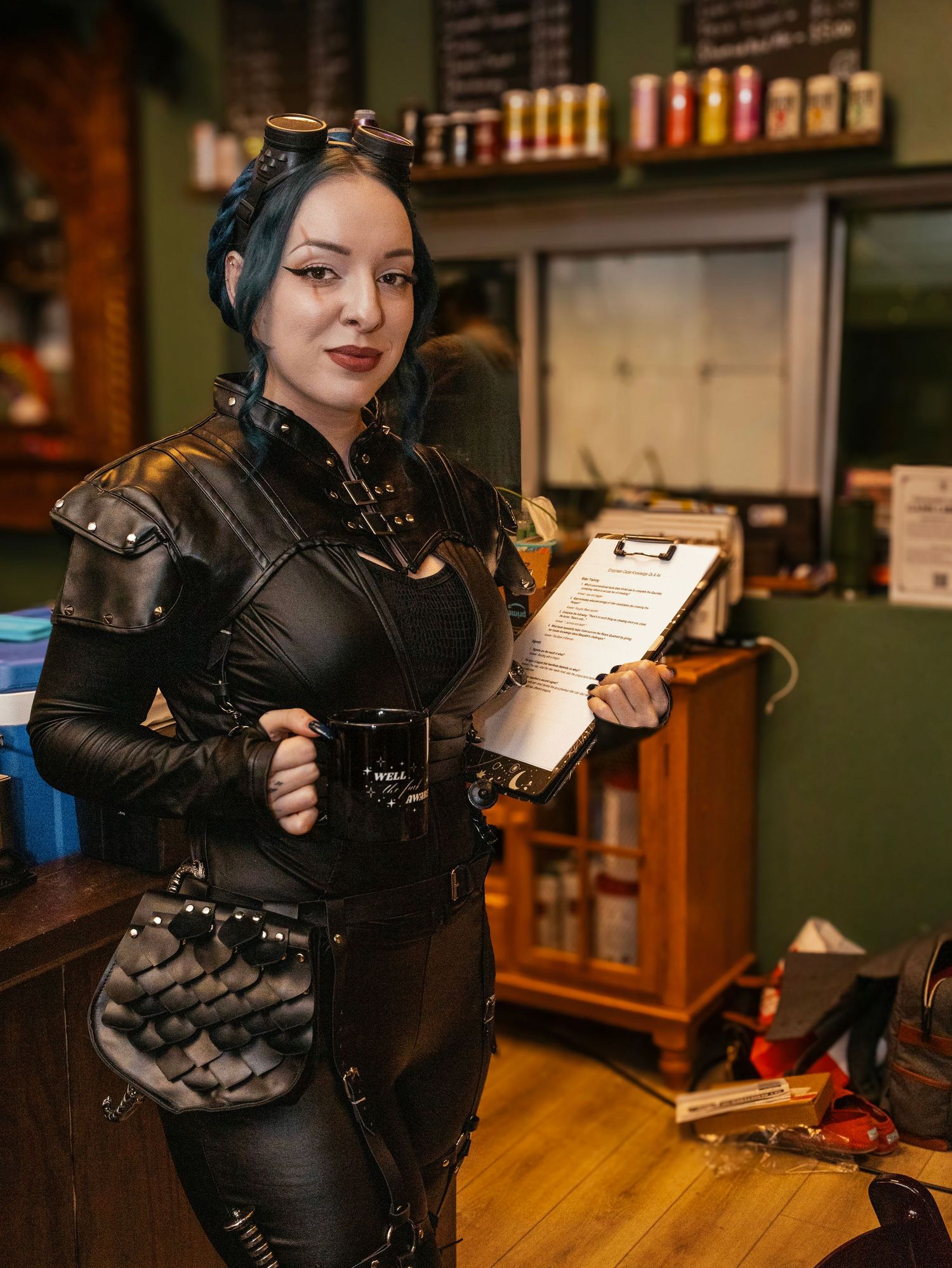 A woman in steampunk leather armor costume holds a clipboard and mug inside a themed cafe setting.