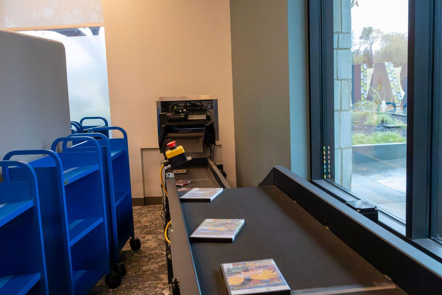 Library checkout counter with blue book carts, receipt printer, and large windows overlooking a sunny outdoor area.