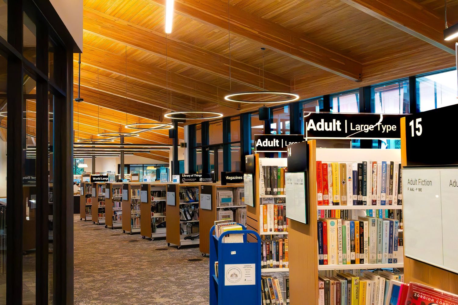 Modern public library interior with wooden ceiling, circular pendant lights, and organized adult book sections.