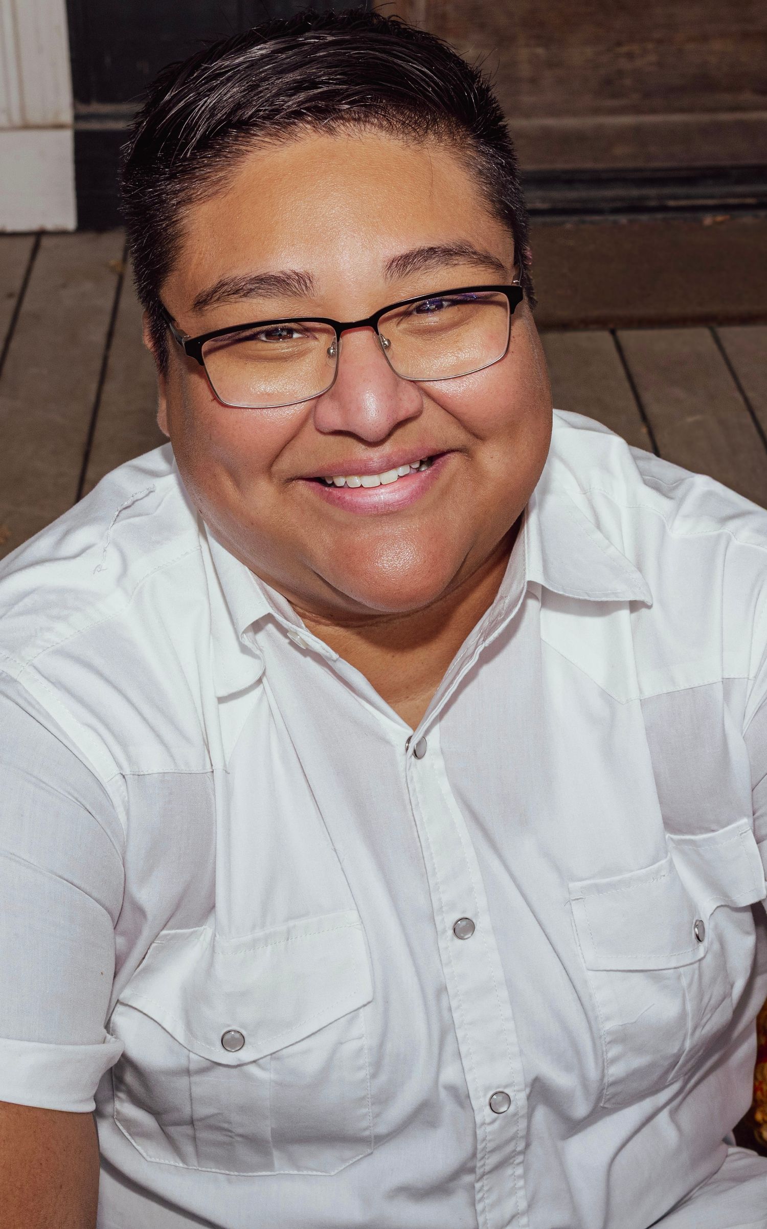 Smiling person with short hair and glasses wearing a white button-up shirt, seated outdoors in a warm, candid portrait.