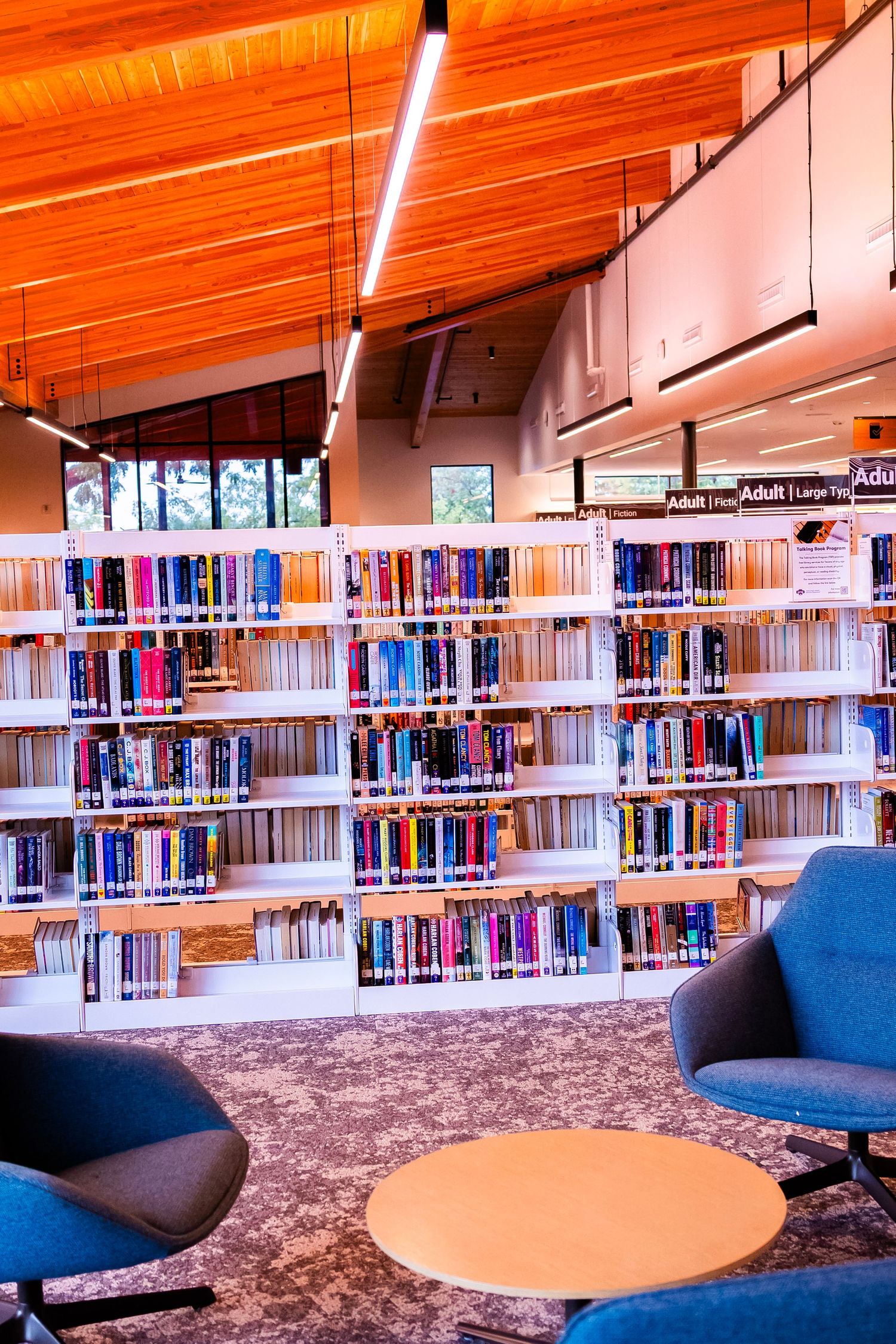 Modern library reading area with colorful bookshelves, blue armchairs, round table, and warm wooden ceiling with bright lighting.