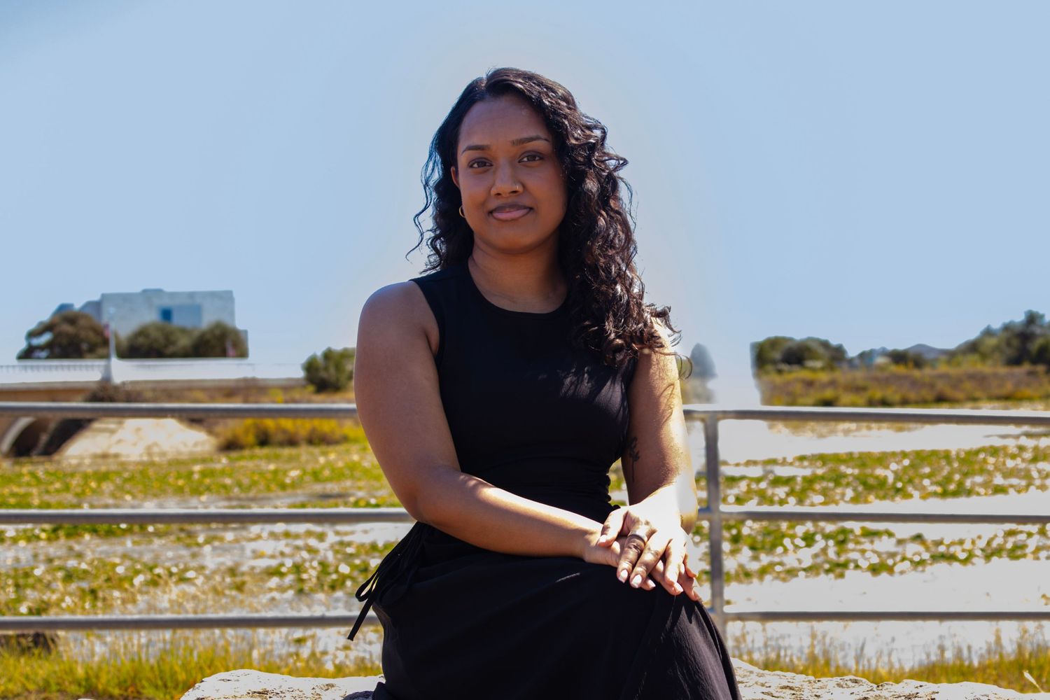 A young woman in a black dress sits outdoors near a fence, with a sunny coastal landscape behind her.