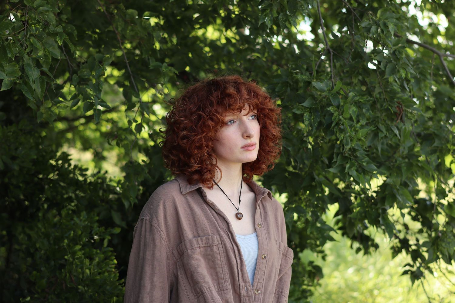 Young woman with curly red hair wearing a brown shirt and pendant necklace standing outdoors among lush green trees.