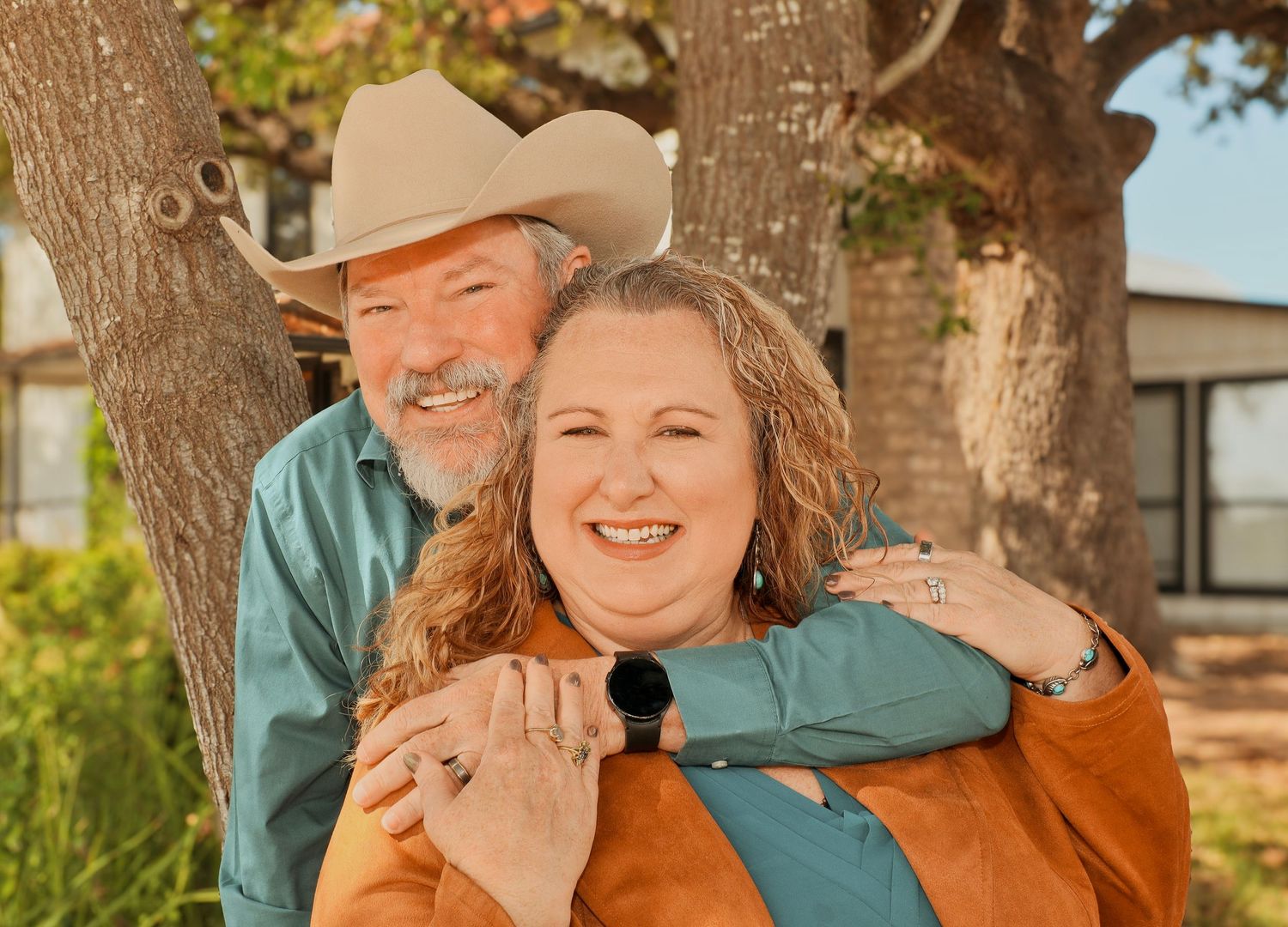 A smiling couple poses outdoors near a tree, man in cowboy hat and teal shirt, woman in orange top.