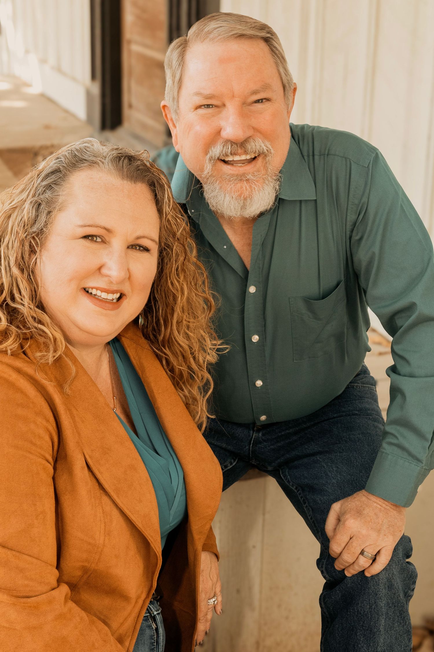 A smiling middle-aged couple posing together, woman in orange jacket with curly hair, man in green shirt with gray beard.