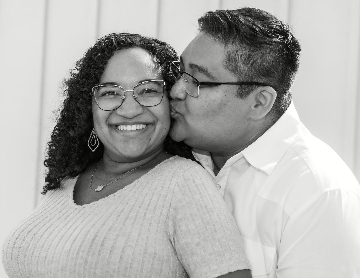 Black and white photo of a smiling woman with curly hair and glasses receiving a kiss on the cheek from a man.