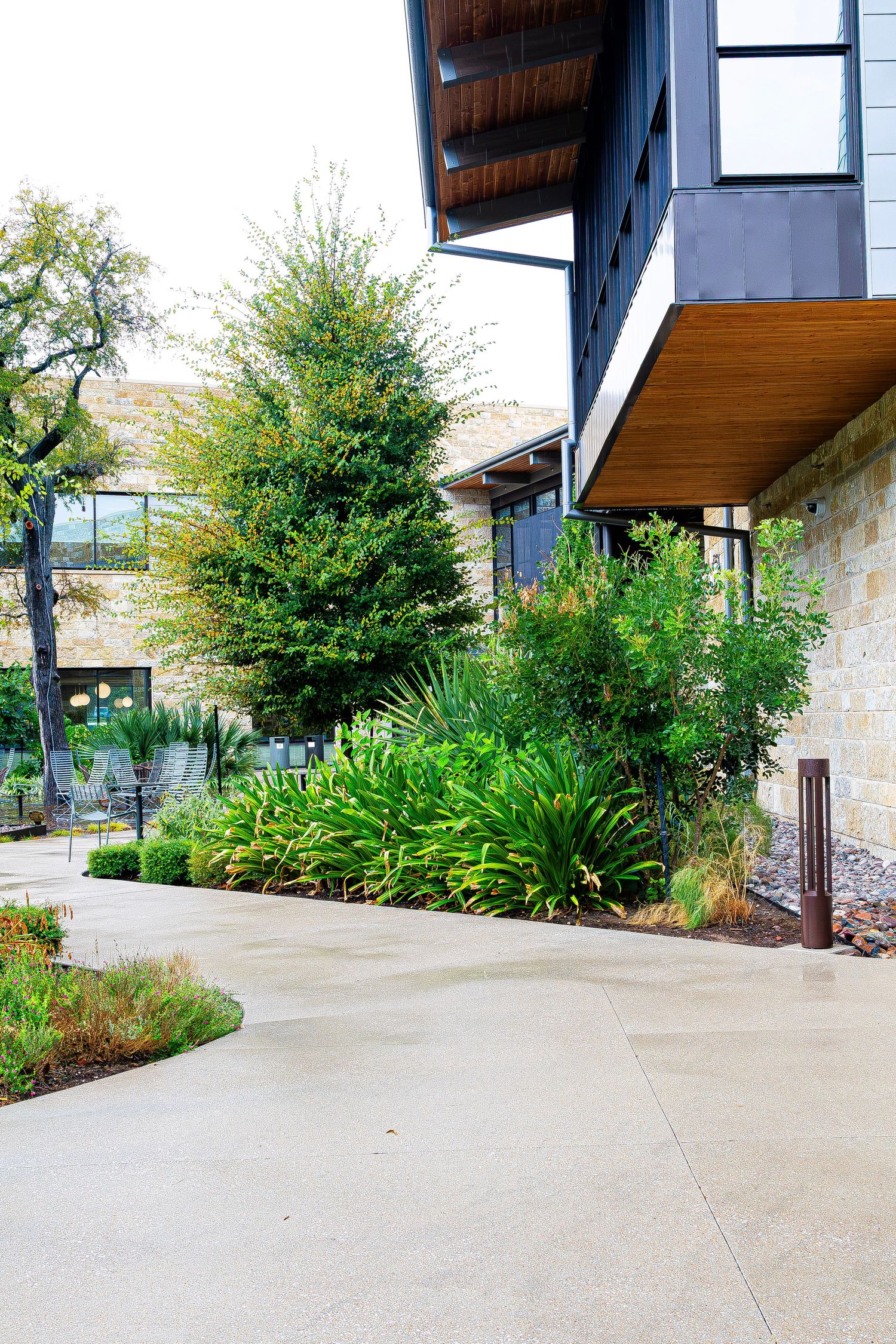 Modern building exterior with lush green landscaping, concrete pathway, and stone facade surrounded by trees.