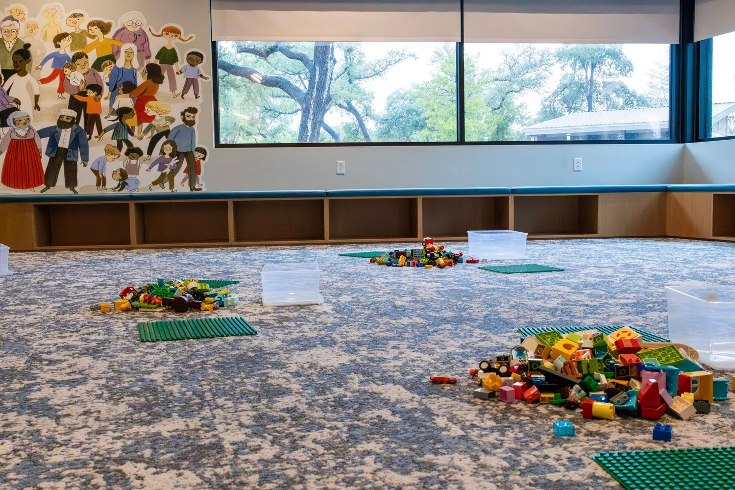 Spacious childrens play area with LEGO building stations on a patterned carpet and a colorful mural by large windows.