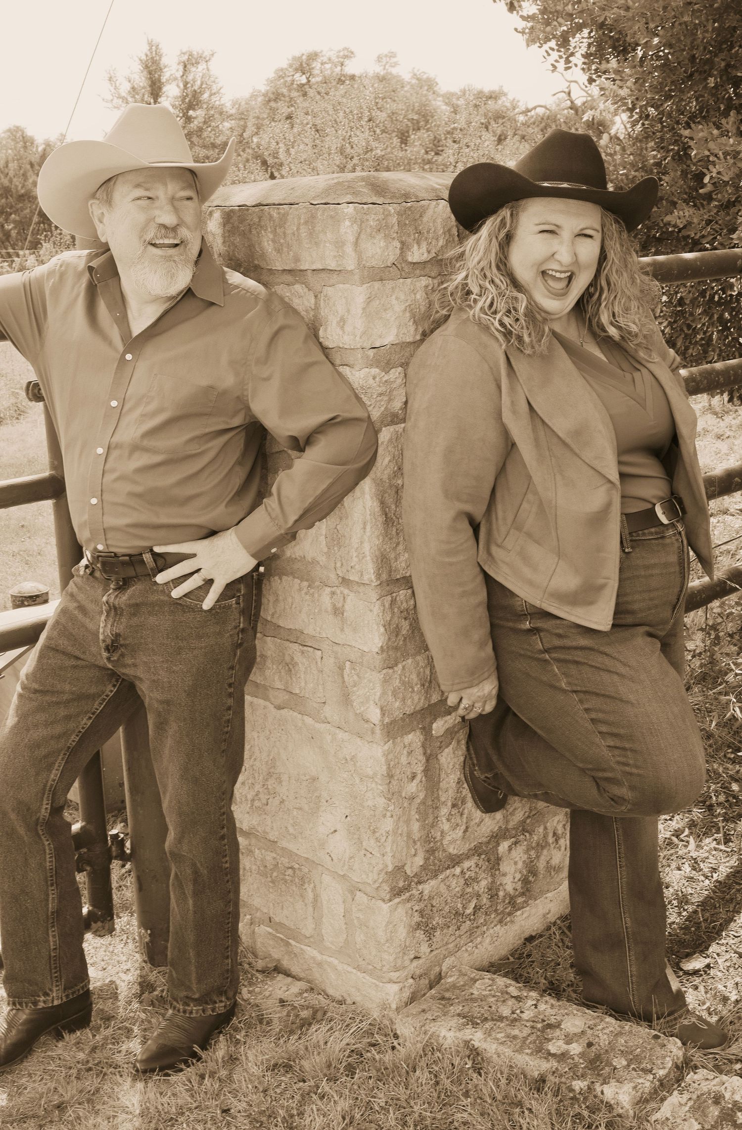 Sepia-toned photo of a smiling man and woman in cowboy hats and jeans leaning against a stone pillar outdoors.
