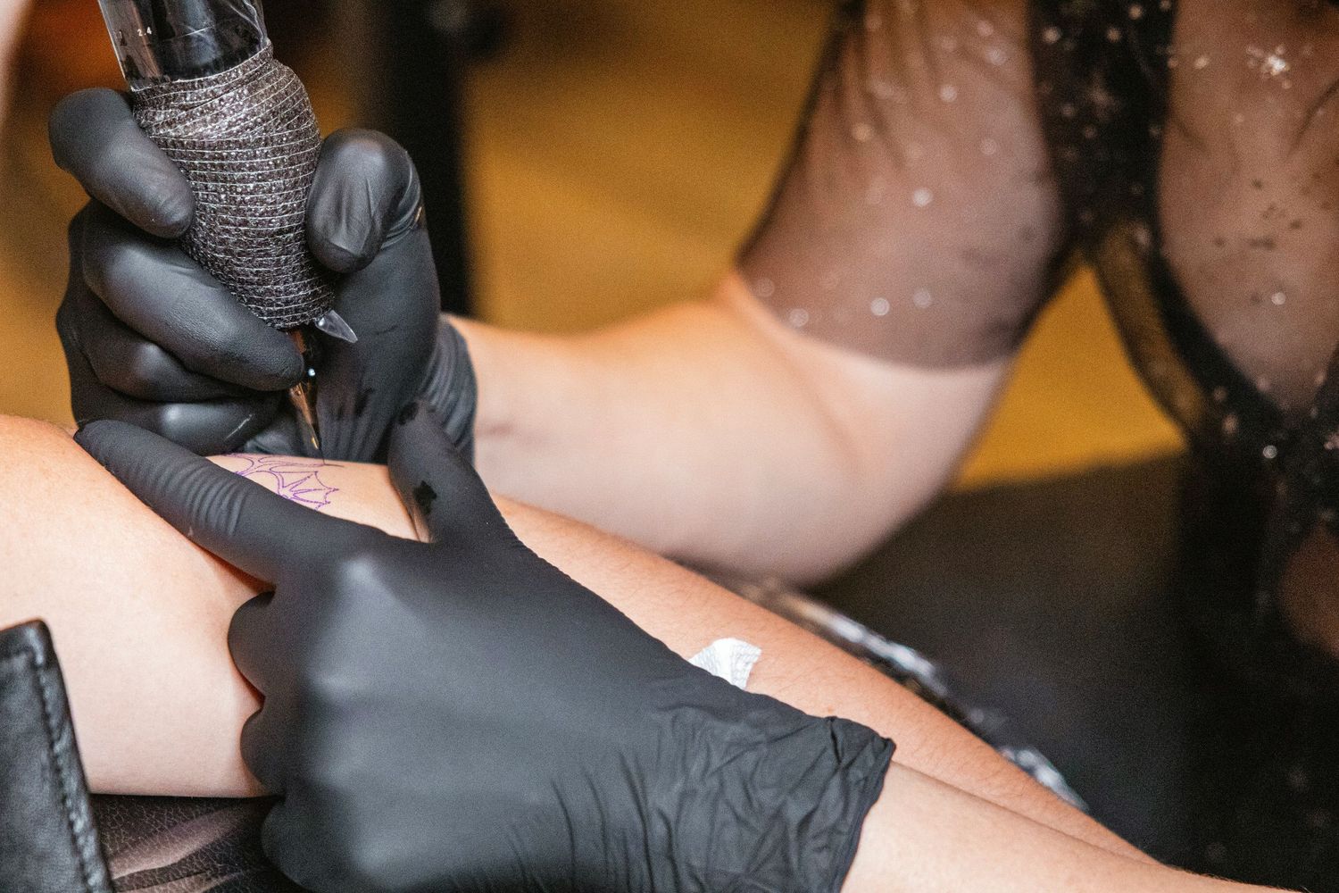 Tattoo artist in black gloves carefully applying a tattoo on a client's leg using a tattoo machine.