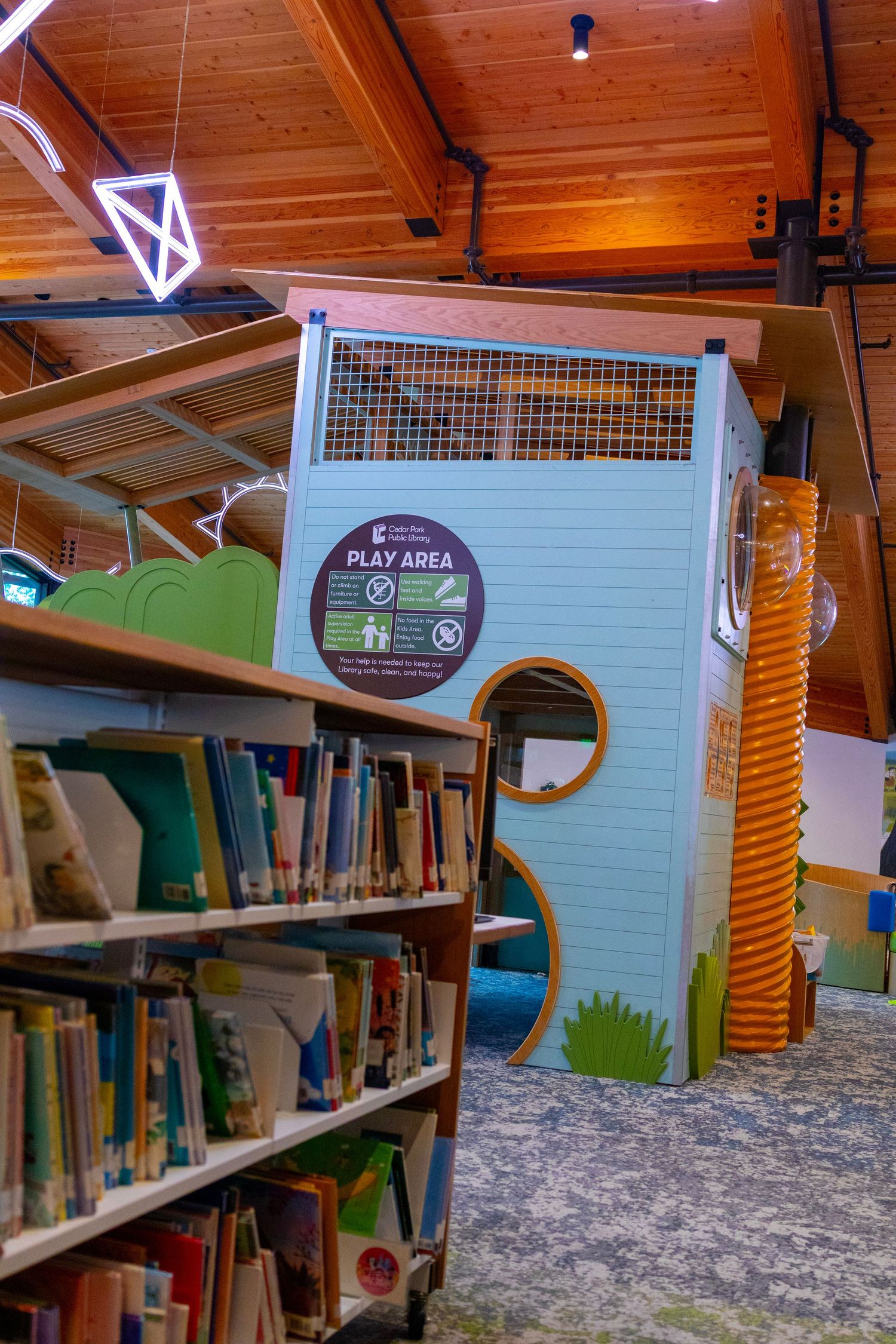 Children's library play area with bookshelf, colorful tunnel slide, and wooden ceiling in a modern public library.