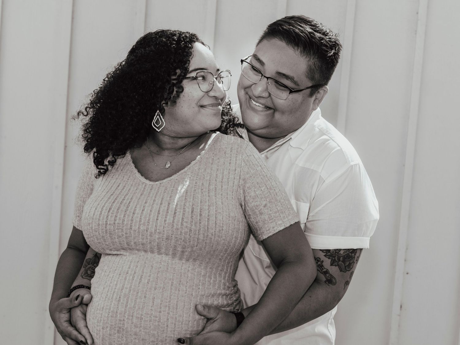 Black and white maternity photo of a smiling couple, pregnant woman in pink sweater embraced by partner outdoors.